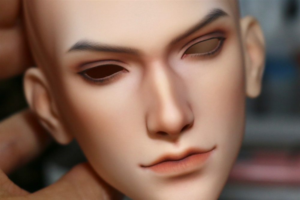 Male doll face-up without eyes yet, with defined dark brows, soft contour and a natural coral lip.