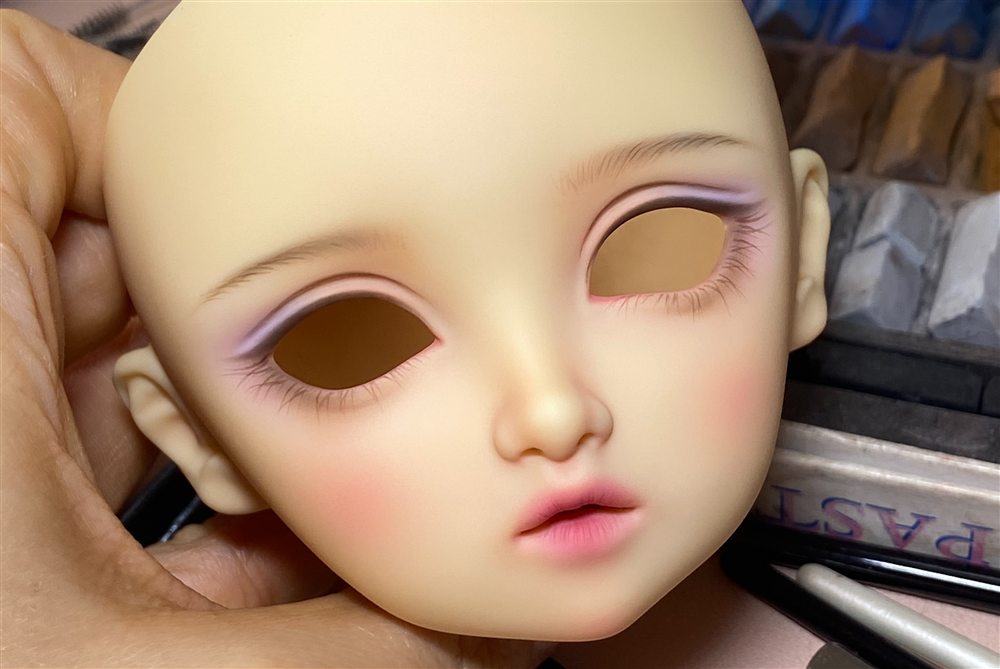 Work in progress: a doll head without eyes, with soft lilac eyeliner, pink blush and a rosy lip, next to pastel pencils.