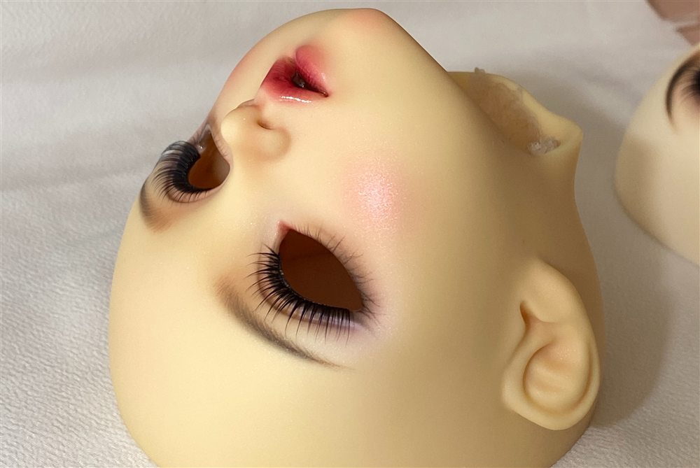 Upside-down view of a doll head with long physical lashes, shimmering blush and a glossy red lip.